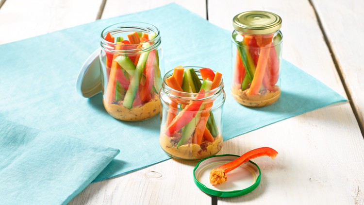 Drei Einmachgläser mit Gemüsesticks aus Paprika und Gurke in Hummus auf einem hellen Holztisch mit türkisfarbenem Tuch.