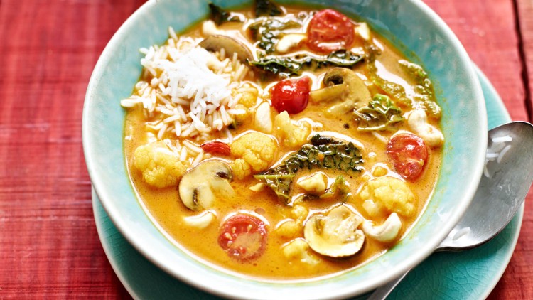 Ein Teller mit gelbem Curry, Reis, Blumenkohl, Tomaten, Champignons, Blattgemüse und Cashewkernen auf einem roten Untergrund.