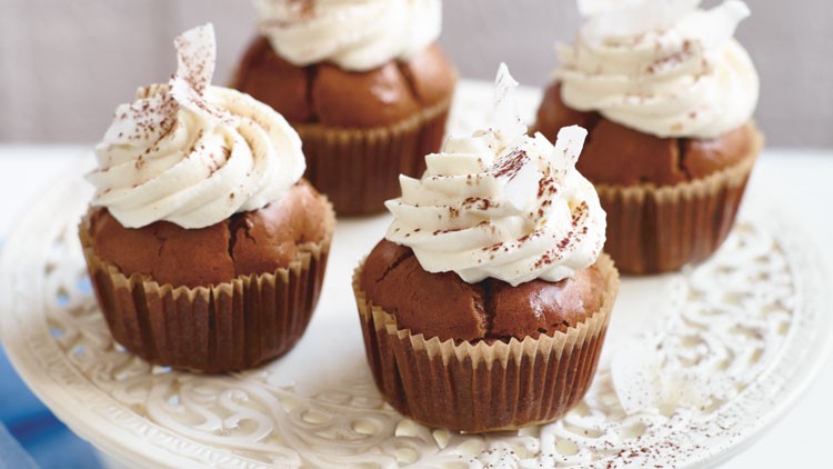 Vier Schokoladenmuffins mit Sahnehaube, Kakaopulver und weißen Dekoelementen auf einem verzierten weißen Tortenständer.