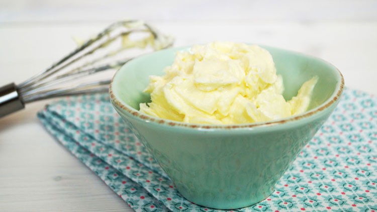 Eine hellgrüne Schüssel mit gelber Creme steht auf einem gemusterten Tuch, im Hintergrund liegt ein Schneebesen.