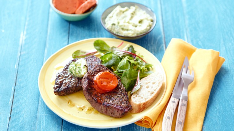 Gegrilltes Steak mit Kräuterbutter, gegrillter Tomate, frischem Salat, Baguettescheibe und Besteck auf gelber Serviette.
