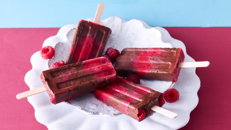Vier rot-braune Eis am Stiel mit Himbeeren auf einem weißen Teller, Hintergrund in Rot und Blau.