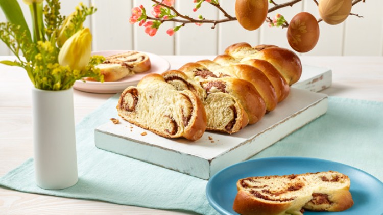 Ein geflochtener Hefezopf mit Füllung liegt in Scheiben geschnitten auf einem Brett, umgeben von Frühlingsdeko und Geschirr.