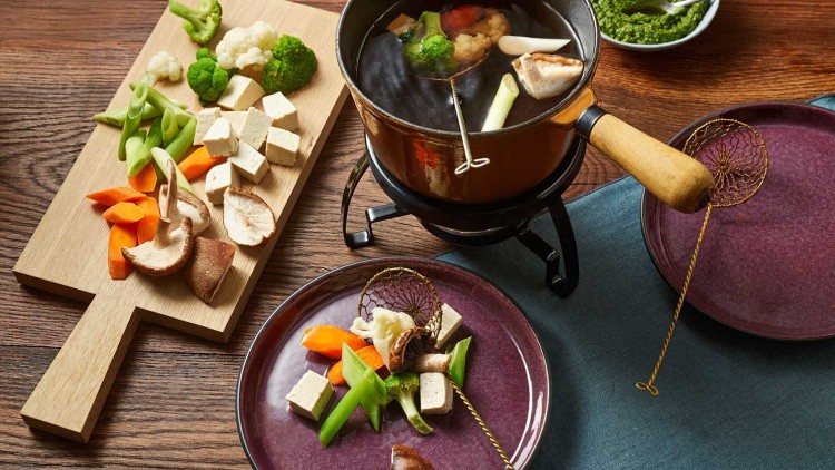 Ein Topf mit Gemüsefondue, einem Holzbrett mit frischem Gemüse und Tofu, zwei Tellern sowie einer Schale mit grünem Dip.