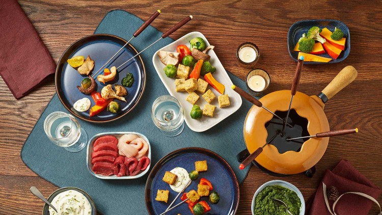 Tisch mit Fondueset, Töpchen mit grüner Soße, Dipp, Gemüsewürfeln und Fleischscheiben, daneben Kerzen und Getränke.