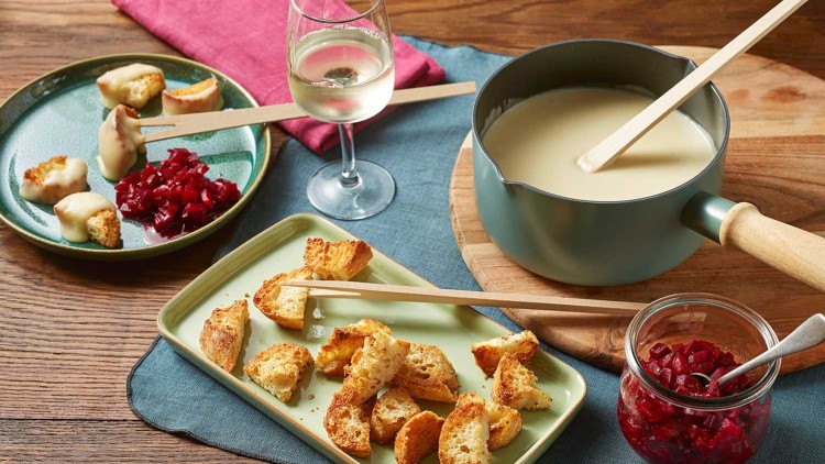 Ein Topf mit geschmolzenem Käse, Brotstücke auf Tellern, Rote-Bete-Würfel im Glas und ein Glas Weißwein auf Holzuntergrund.