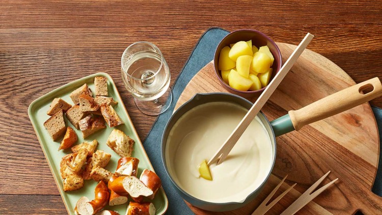 Auf einem Holztisch stehen ein Fonduetopf mit Käsecreme, Brotstücke, Kartoffelwürfel, Holzspieße und ein Glas Weißwein.