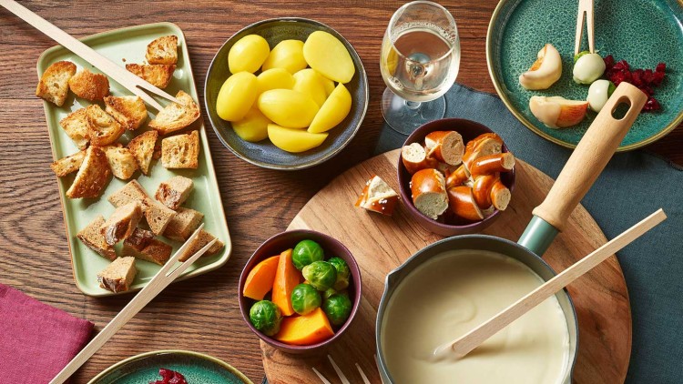 Tisch mit Fonduetopf, Brotstücken, Kartoffeln, Gemüse wie Rosenkohl und Kürbis, Brezeln, Käse und Glas Weißwein.