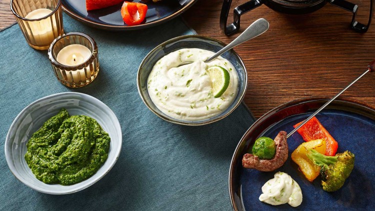 Fondue mit Gemüse und Fleisch auf blauen Tellern, dazu Dips in Schalen und zwei brennende Kerzen auf Holztisch mit Stofftuch.