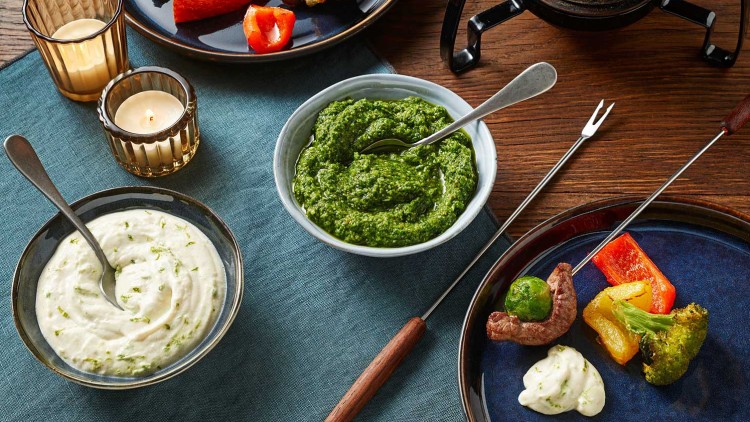 Zwei Teller mit gegrilltem Gemüse und Fleisch, Fonduespieße, zwei Dippschalen und Teelichter auf Holztisch und Stofftuch.
