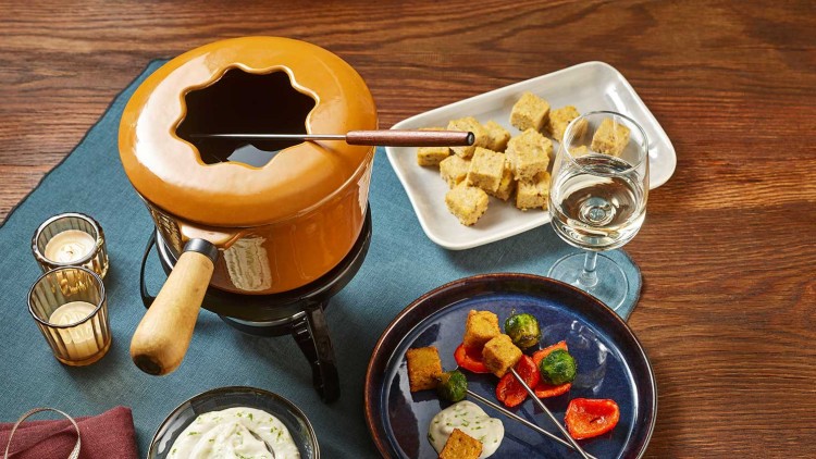 Ein orangefarbener Fonduetopf mit Holzgriff, Gemüsespieße auf einem Teller und Dipschale auf einem gedeckten Tisch.