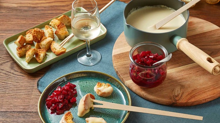 Brotscheiben, Käsefondue, rote Beilagen in Glas und auf Teller, Weißweinglas, Holzlöffel und blaue Tischdecke auf Holztisch.