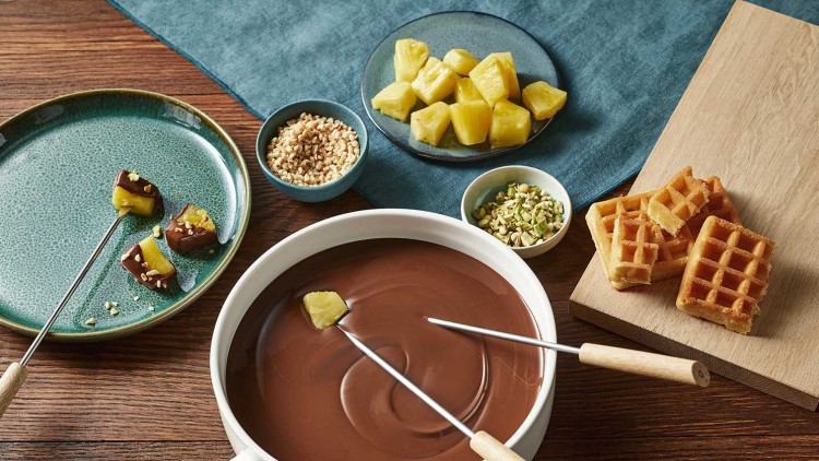 Karamellisierte Ananasstücke, Waffeln und Toppings um eine weiße Fondue-Schale mit flüssiger Schokolade und zwei Fondue-Gabeln.