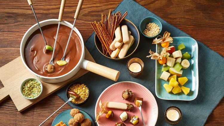 Ein Schokoladenfondue mit Früchten, Gebäck, Waffeln und Schälchen mit Nüssen auf einem Holz- und Stoffuntergrund.