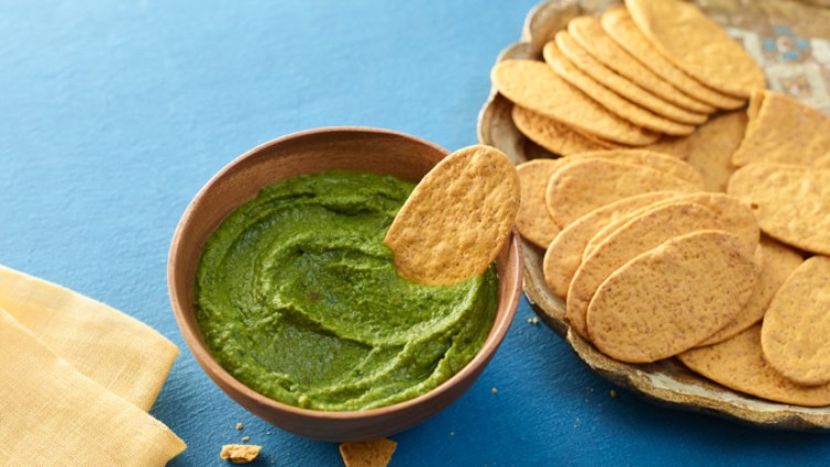 Eine Holzschale mit grüner Creme und ein angelehnter Chip, daneben ein Teller mit weiteren Chips auf blauem Untergrund.