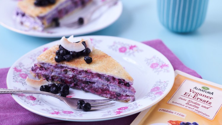 Ein Stück Beeren-Crepe-Kuchen mit Kokosflocken und Heidelbeeren auf einem Blumenteller, daneben Alnatura Veganer Ei-Ersatz.
