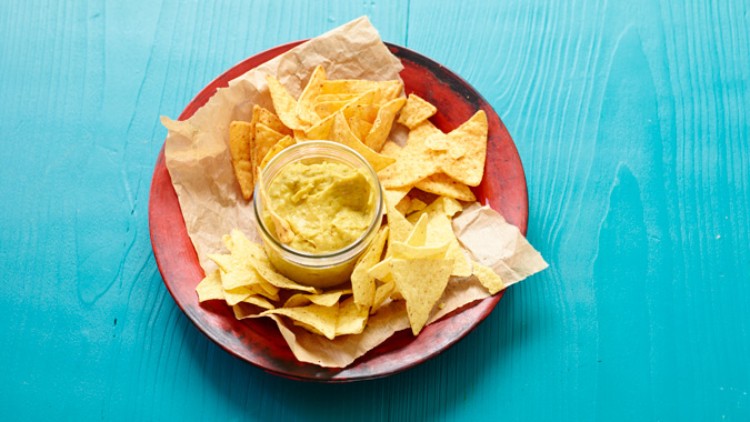 Ein roter Teller mit gelben Tortillachips und einem Glas Guacamole steht auf einem türkisfarbenen Holztisch.