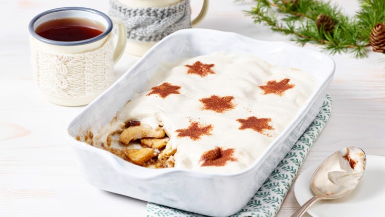 Eine weiße Auflaufform mit Schichtdessert und Zimtsternen auf Sahne, daneben Tee in einer Tasse mit gestrickter Hülle.