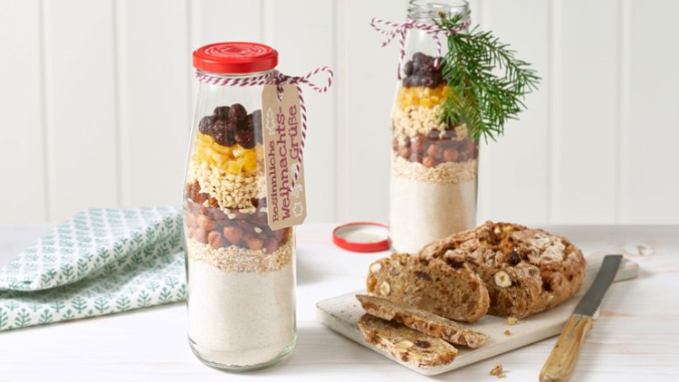 Zwei Glasflaschen mit Backmischungen, Dekoration und Anhänger stehen neben angeschnittenem Nussbrot auf einem hellen Tisch.