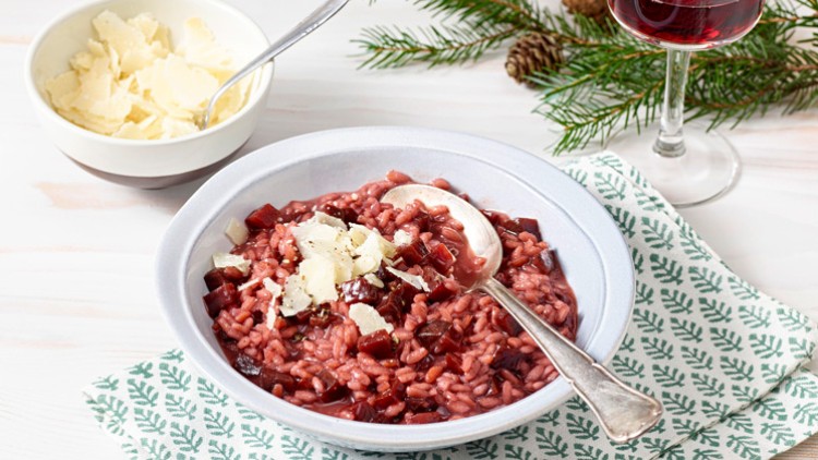 Rosa Rote-Bete-Risotto mit Käsespänen in einer weißen Schale, daneben ein Glas Rotwein und ein Zweig Tannengrün.