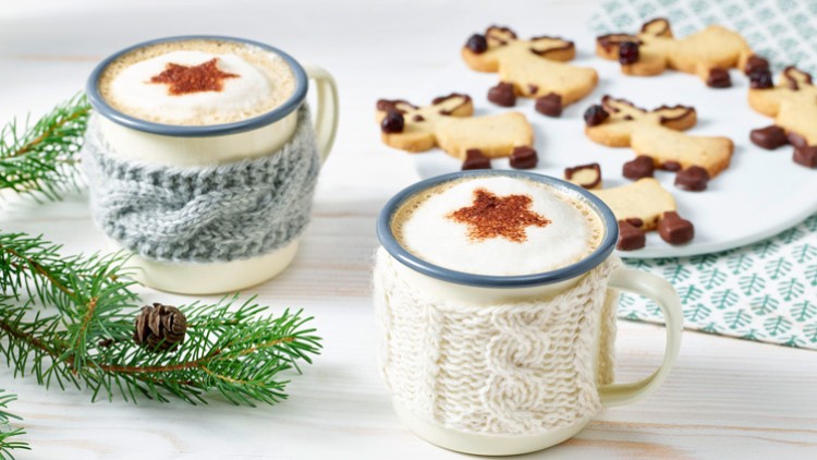 Zwei Tassen mit Milchschaum und Stern-Dekor in gestrickten Hüllen stehen neben Tannenzweig und Rentier-Keksen auf hellem Tisch.