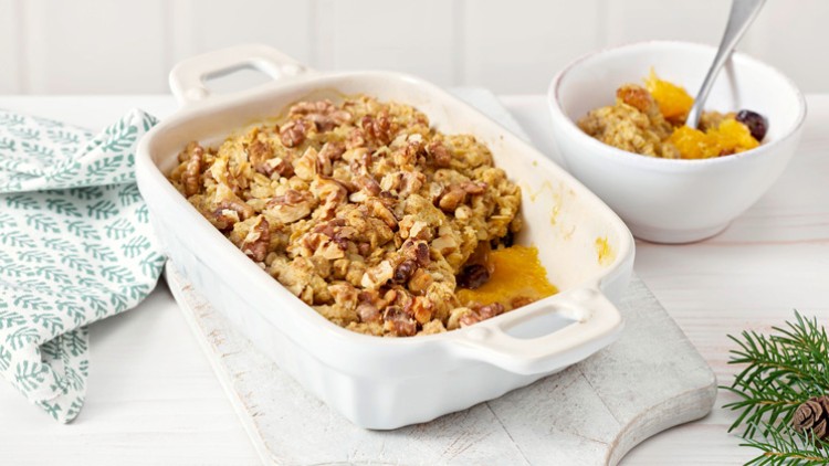 Auf einer weißen Auflaufform liegt ein Walnussauflauf, daneben steht eine Schüssel mit Obst und ein Löffel auf hellem Holztisch.