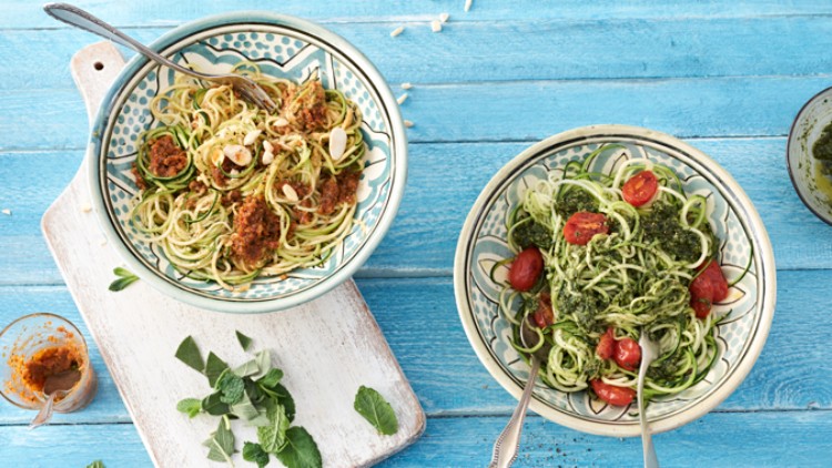 Zwei Schalen mit Zucchininudeln, einmal mit rotem Pesto und Mandeln, einmal mit grünem Pesto und Tomaten, auf blauem Holztisch.