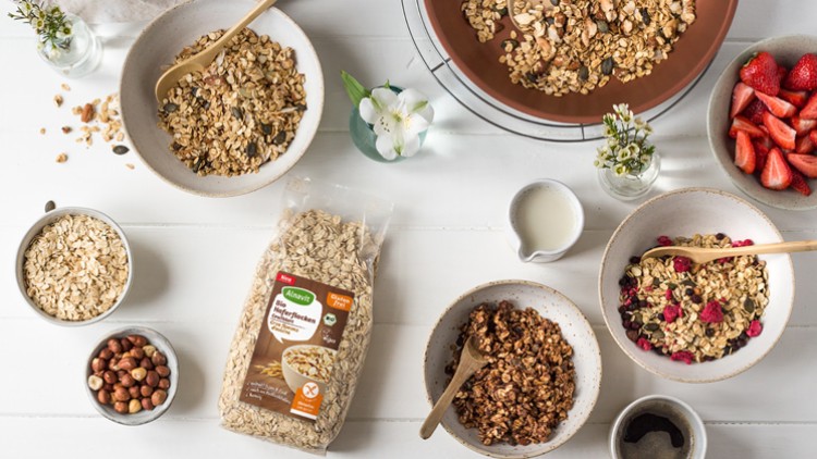 Mehrere Schalen mit Haferflocken, Müsli, frischen Erdbeeren, gerösteten Nüssen, einer Milchkanne und einer Alnatura-Haferflockenpackung.