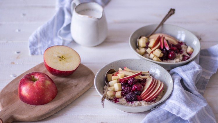 Zwei Schalen mit Porridge, Apfelstücken und Beeren sowie ein Holzbrett mit einem roten Apfel vor einer Kanne Milch.