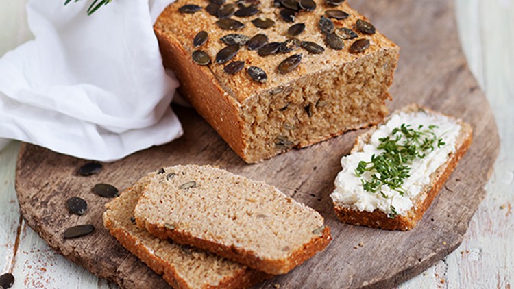 Ganze Kastenbrot aus Vollkorn mit Kürbiskernen, zwei Scheiben Brot und eine bestrichene Scheibe mit Frischkäse und Kräutern.