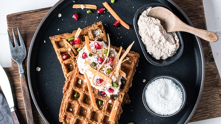 Zwei Waffeln auf einem schwarzen Teller, belegt mit Creme, Granatapfelkernen und Pistazien, daneben Schälchen mit Creme und Puderzucker.