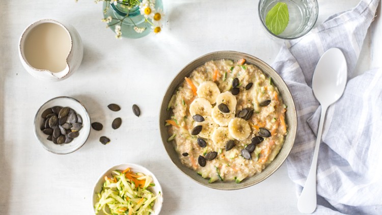 Eine Schüssel Porridge mit Bananenscheiben, geraspeltem Gemüse und Kürbiskernen auf einem weißen Tisch mit weiteren Zutaten.