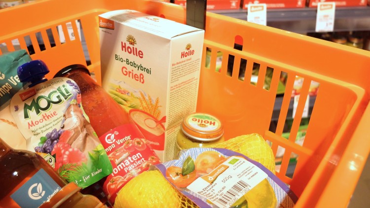 Orange Einkaufskorb mit Bio-Produkten wie Babybrei, Smoothie, passierte Tomaten und Zitronen, im Hintergrund Regale mit Babygläschen.