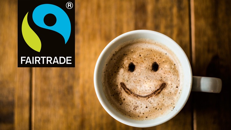 Tasse Kaffee mit Schaum und einem lächelnden Gesicht aus Kakaopulver, links oben das Fairtrade-Logo auf holzfarbenem Hintergrund.