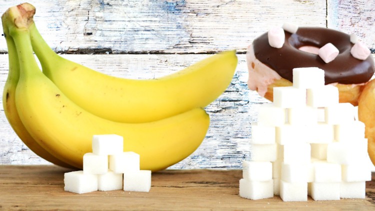 Roter Apfel, grüner Apfel, Bananen, Schokodonut, Karamelldonut und Würfelzucker auf einem Holztisch vor weißer Wand.