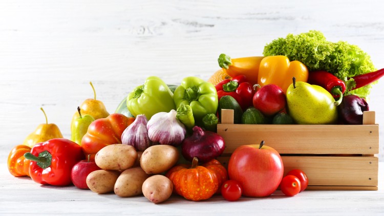 Verschiedene Obst- und Gemüsesorten wie Paprika, Äpfel, Kartoffeln, Knoblauch und Salat neben einer Holzkiste auf hellem Hintergrund.