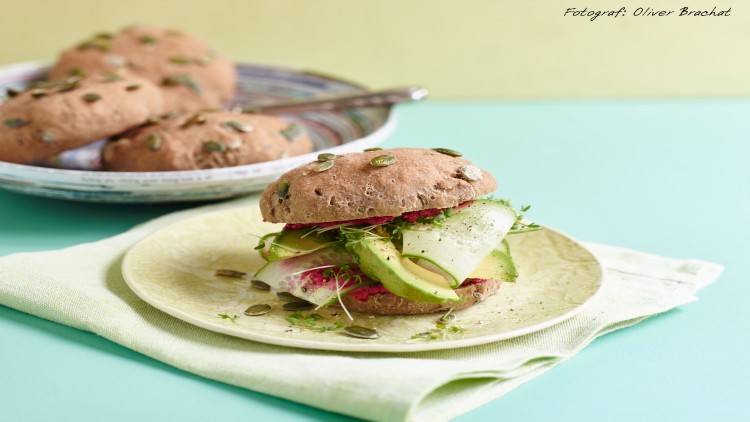 Vollkornbrötchen mit Kürbiskernen, gefüllt mit Avocado, Roter Bete-Creme, Zucchinistreifen und frischen Kräutern auf einem Teller.