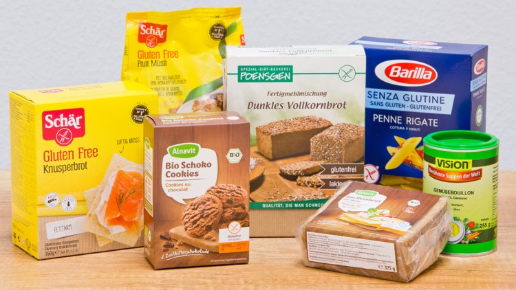 Verschiedene glutenfreie Lebensmittel wie Brot, Kekse, Pasta, Müsli und Gemüsebouillon stehen auf einer Holzfläche.