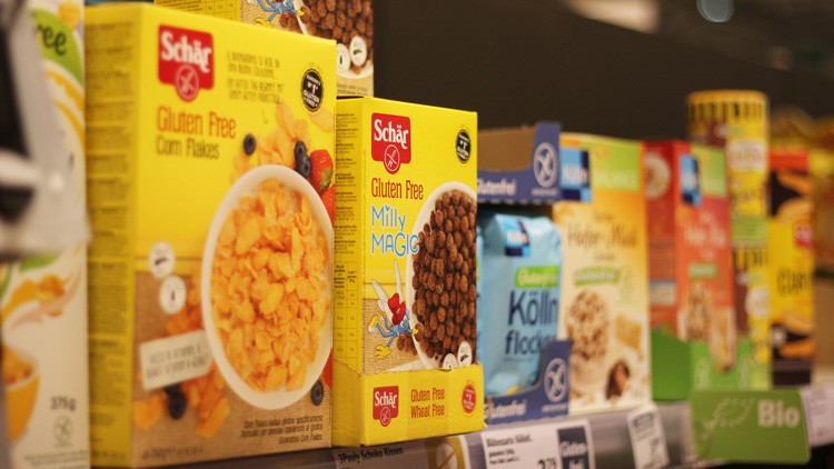 Glutenfreie Frühstückscerealien verschiedener Marken, darunter Schär Corn Flakes und Milly Magic, stehen in Regalreihen.