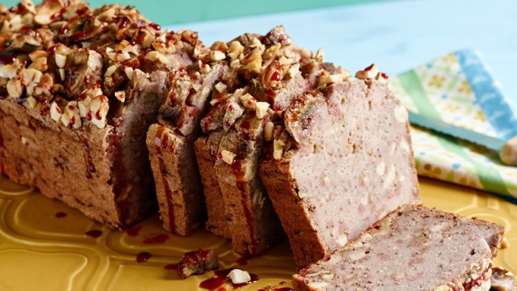 Ein angeschnittenes Bananenbrot mit Nüssen und Fruchtsoße liegt auf gelber Platte, daneben ein Messer auf buntem Stoff.