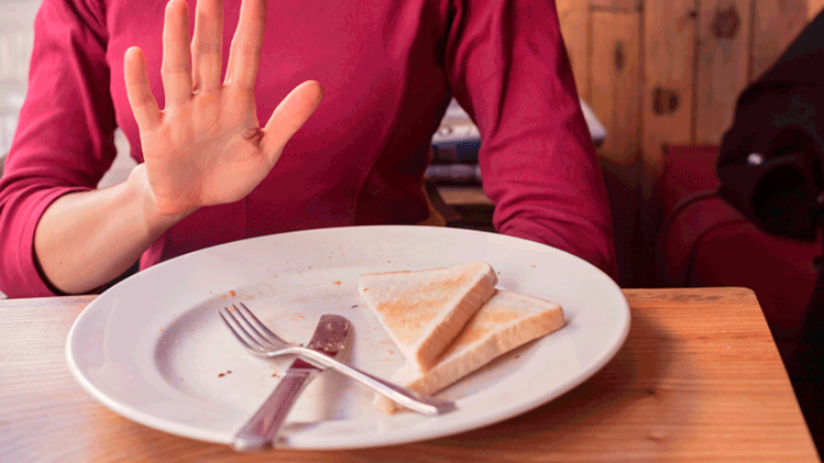 Eine Person in rotem Oberteil hebt die Hand, vor ihr steht ein Teller mit zwei Toaststücken, Messer und Gabel.