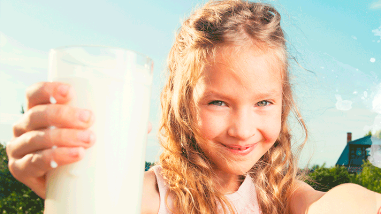 Kind mit langen lockigen Haaren hält ein gefülltes Glas Milch in der Hand, Hintergrund ist ein sonniger Außenbereich.