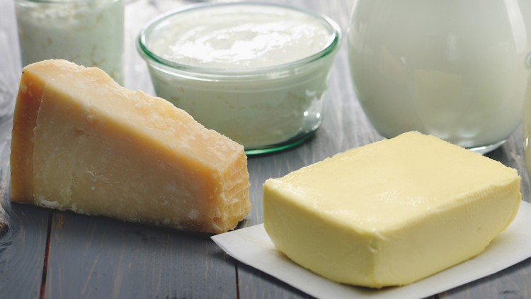 Ein Stück Hartkäse, ein Stück Butter auf Papier und Gläser mit Milchprodukten stehen auf einer grauen Holzfläche.
