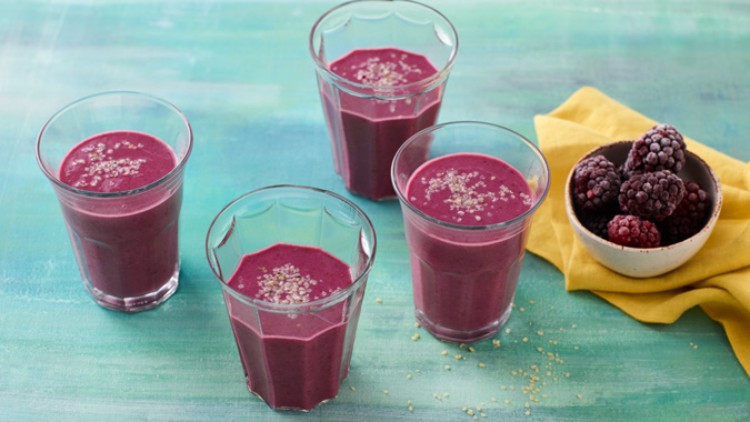 Vier Gläser mit lila Smoothie, bestreut mit Samen, stehen auf grünem Untergrund, daneben eine Schale gefrorene Beeren.