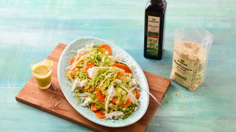 Bunter Salat aus Karotten, Kraut und Feta auf einer ovalen Platte, daneben Hanföl, Hanfsamen und ein Glas Dressing.