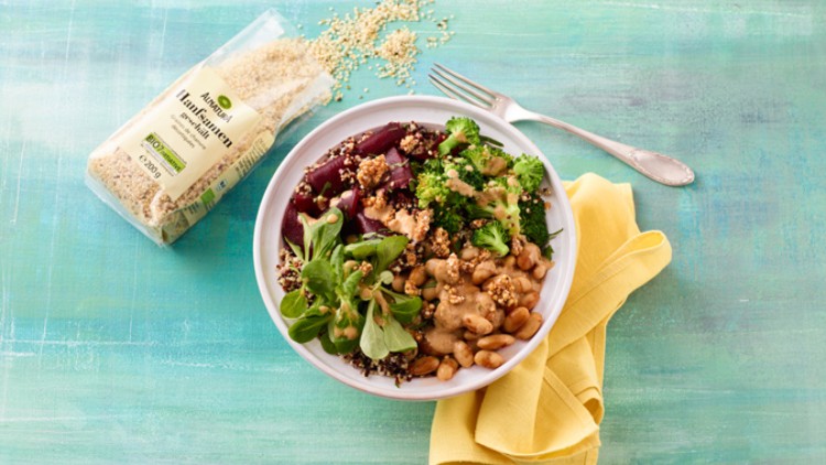 Bunte Bowl mit Quinoa, Brokkoli, Rote Bete, Bohnen, Feldsalat und Dressing, daneben eine offene Packung Quinoa und gelbe Serviette.