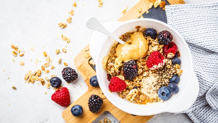 Weiße Schüssel mit Müsli, Beeren, Erdnussbutter und Samen auf einem Holzbrett, daneben ein Stofftuch und verstreute Zutaten.