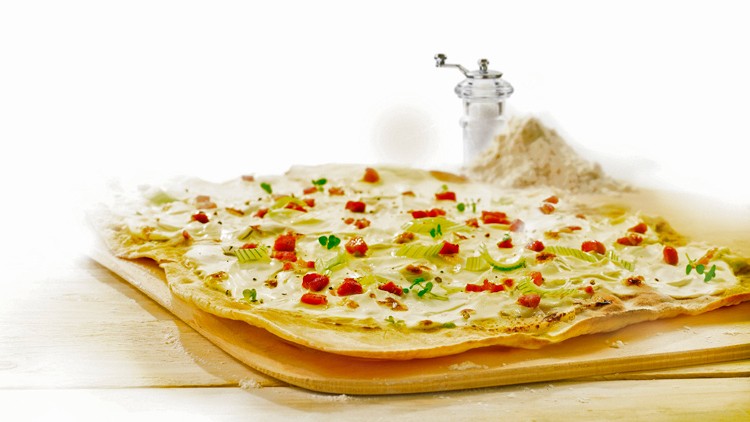 Ein rechteckiger Flammkuchen mit Schmand, grünen Lauchringen, Speckwürfeln und Kresse auf einem Holzbrett, daneben Mehl und Pfeffer.