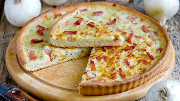 Ein Zwiebelkuchen mit knusprigem Rand, Speckwürfeln und Käse auf einem Holzbrett, umgeben von weißen Zwiebeln.