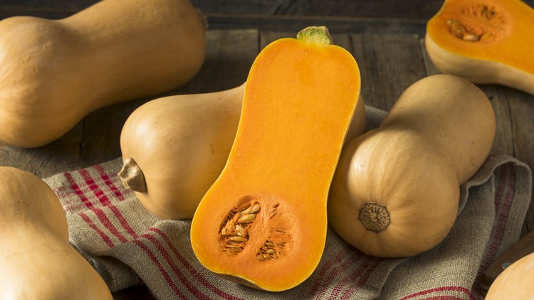 Mehrere Butternut-Kürbisse liegen auf einem Holztisch, einer davon ist längs halbiert und zeigt orangefarbenes Fruchtfleisch mit Kernen.